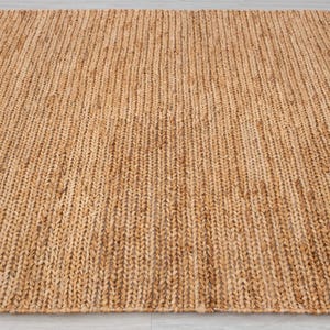 May include: A rectangular, woven jute rug in a natural tan color. The rug has a textured surface with vertical ribbing, creating a simple, yet elegant design. This floor covering is suitable for various interior styles.