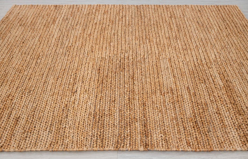 May include: A rectangular, woven jute rug in a natural tan color. The rug has a textured surface with vertical ribbing, creating a simple, yet elegant design. This floor covering is suitable for various interior styles.
