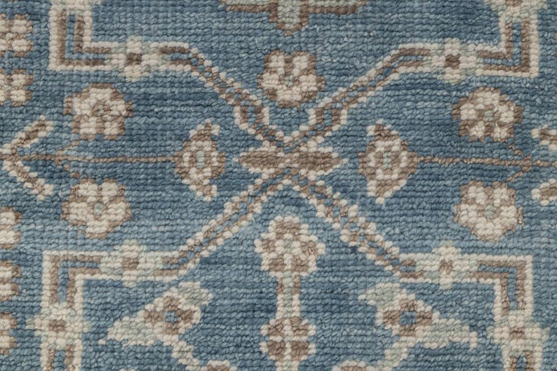 May include: Close-up of a blue rug with a detailed pattern. The design features repeating floral and geometric shapes in shades of beige and brown, creating a textured surface. The rug's color palette is a mix of blue, beige, and brown.