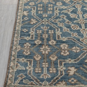 May include: A section of a blue area rug with an intricate geometric pattern. The rug features a central field of blue with beige and brown accents. The border is a darker brown with a fringe along the edge. The rug is on a light gray surface.