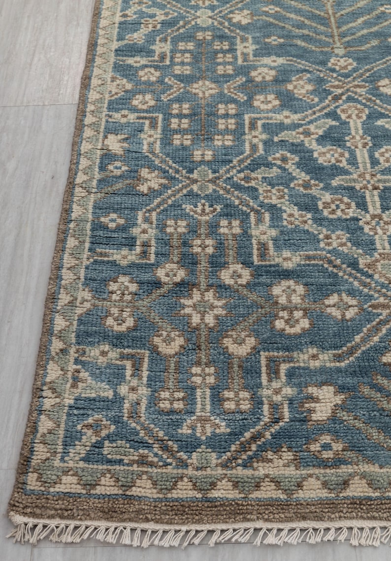 May include: A section of a blue area rug with an intricate geometric pattern. The rug features a central field of blue with beige and brown accents. The border is a darker brown with a fringe along the edge. The rug is on a light gray surface.