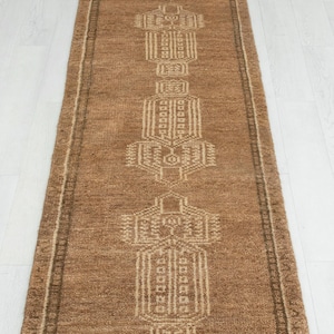 Könnte beinhalten: Ein langer, rechteckiger Läufer in mittlerem Braunton mit einer Reihe sich wiederholender, symmetrischer geometrischer Muster in einem helleren Beige. Der Teppich hat einen strukturierten, gewebten Flor und eine kurze, weiße Fransenborte.