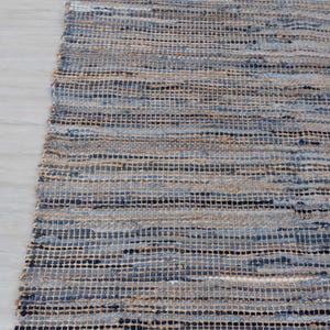 May include: A rectangular rug with a woven design. The rug features a mix of blue, beige, and black horizontal stripes. The texture appears to be made of natural fibers, creating a casual and inviting aesthetic. The rug is placed on a light-colored wooden floor.