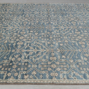 May include: A large, rectangular rug with a detailed, repeating pattern. The rug features a blue background with a beige and gray floral design. The border is a lighter beige color, and the rug is placed on a light gray surface.