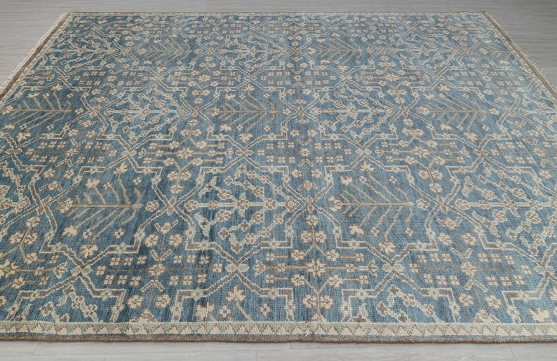 May include: A large, rectangular rug with a detailed, repeating pattern. The rug features a blue background with a beige and gray floral design. The border is a lighter beige color, and the rug is placed on a light gray surface.
