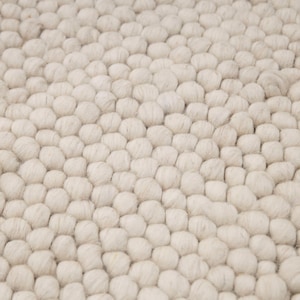 May include: Close-up of a cream-colored rug with a textured, bubble-like pattern. The rug's surface is densely covered with rounded, wool-like elements, creating a soft, tactile appearance. The neutral color and unique texture suggest a cozy, modern home decor item.
