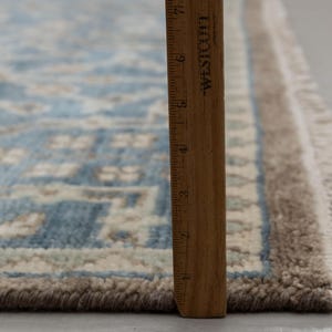 May include: A section of a blue and beige patterned rug with a fringed edge. A wooden ruler is placed vertically along the rug's edge, showing measurements in inches. The rug has a detailed, geometric design.