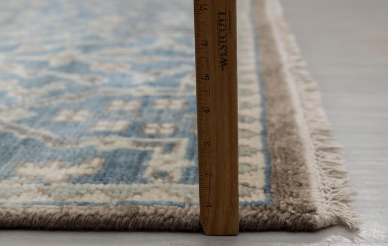 May include: A section of a blue and beige patterned rug with a fringed edge. A wooden ruler is placed vertically along the rug's edge, showing measurements in inches. The rug has a detailed, geometric design.