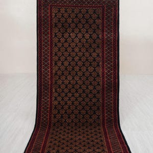 Puede incluir: Una alfombra rectangular con un fondo azul marino oscuro y un patrón repetido de formas florales marrones. La alfombra tiene un borde rojo con un diseño geométrico y bordes con flecos. La alfombra se exhibe sobre una superficie de color claro.