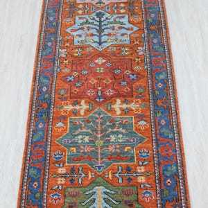 Puede incluir: Una alfombra pasillera rectangular larga con un fondo naranja vibrante y una serie de medallones geométricos repetitivos en azul, verde, amarillo y rosa. Cada medallón contiene motivos de árboles estilizados y patrones florales.