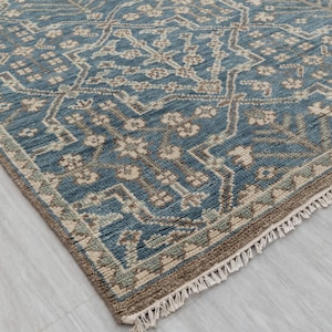 May include: A close-up of a blue and beige patterned rug. The rug features a detailed geometric design with floral elements. The edges are finished with a fringe. The rug is on a light-colored surface.