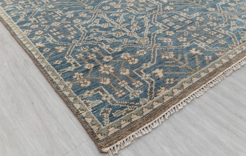 May include: A close-up of a blue and beige patterned rug. The rug features a detailed geometric design with floral elements. The edges are finished with a fringe. The rug is on a light-colored surface.