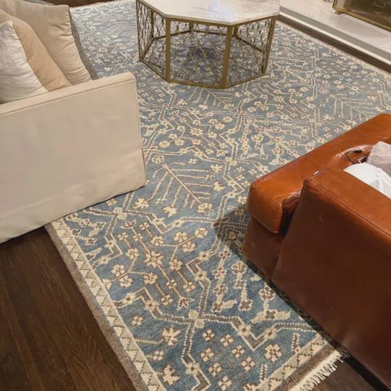 May include: A blue and beige patterned area rug with a distressed look. The rug is rectangular and features a repeating floral and geometric design. The rug is placed on a dark wood floor, with a white sofa and a brown leather chair visible.