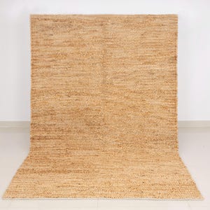 May include: A large, rectangular jute rug with a natural, light brown color. The rug has a woven texture and is displayed against a white wall and floor. The rug is rolled up at the bottom.