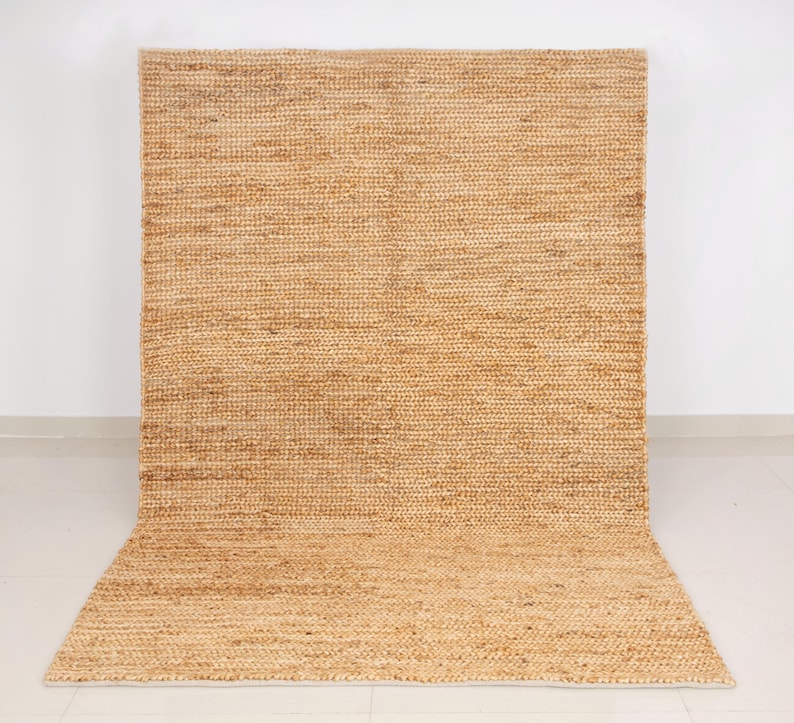 May include: A large, rectangular jute rug with a natural, light brown color. The rug has a woven texture and is displayed against a white wall and floor. The rug is rolled up at the bottom.