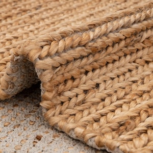 May include: Close-up of a natural fiber rug with a woven, braided texture. The rug is a light brown color, with a rolled-up edge revealing the underside. The weave is tight and uniform, creating a durable and textured surface.