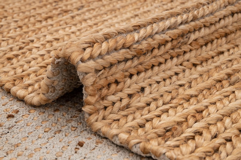 May include: Close-up of a natural fiber rug with a woven, braided texture. The rug is a light brown color, with a rolled-up edge revealing the underside. The weave is tight and uniform, creating a durable and textured surface.