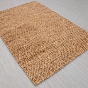 May include: A rectangular, woven jute rug in a natural tan color. The rug has a textured surface with a ribbed pattern, suitable for indoor use. The rug is placed on a light gray surface.