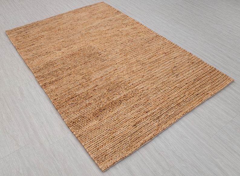 May include: A rectangular, woven jute rug in a natural tan color. The rug has a textured surface with a ribbed pattern, suitable for indoor use. The rug is placed on a light gray surface.