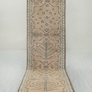 May include: A long rectangular runner rug featuring a soft peach background with a repeating pattern of stylised trees and floral motifs in shades of dark brown and light blue. The rug is framed by a geometric border and has white fringe ends.