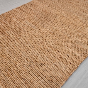 May include: A rectangular, woven jute rug in a natural tan color. The rug has a textured surface with a diagonal pattern, suitable for indoor use. The rug is made of natural fibers.