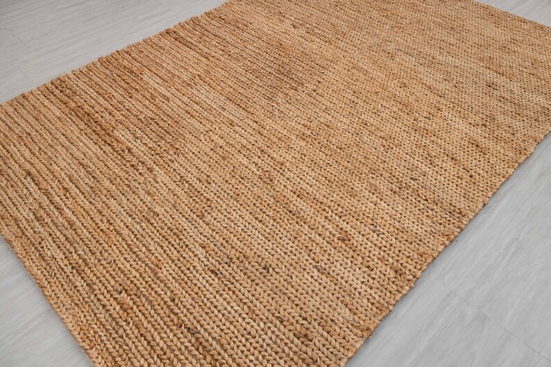 May include: A rectangular, woven jute rug in a natural tan color. The rug has a textured surface with a diagonal pattern, suitable for indoor use. The rug is made of natural fibers.