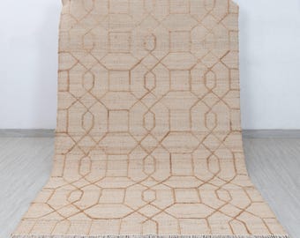 May include: Beige jute rug with a geometric pattern. The rug features a repeating design of interconnected shapes in a lighter shade of beige. The rug has a fringed edge and is displayed against a white wall and light gray floor.