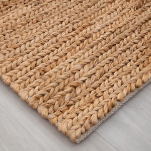 May include: Close-up of a natural fiber rug with a braided design. The rug is made of a light brown material and has a textured surface. The rug's edge is visible, showing a lighter-colored border.