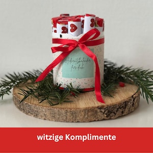 Könnte beinhalten: Ein zylindrischer Geschenkbehälter mit rotem Band und dem Etikett „Adventskalender für dich“. Er ist gefüllt mit kleinen, verpackten Geschenken und steht auf einer Holzscheibe mit Grünzeug. Der Bildgrund trägt den Text „witzige Komplimente“.