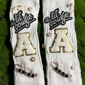 May include: A pair of white ribbed socks with the text "a lil bougie" in black and white script. Each sock features a large, fluffy letter "A" with gold trim, decorative pins, and beaded accents.