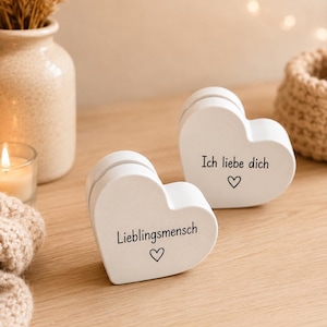 Può includere: Due blocchi di legno bianchi a forma di cuore con testo nero e una piccola icona a forma di cuore. Un blocco mostra il testo tedesco 'Lieblingsmensch' e l'altro 'Ich liebe dich'. Ogni cuore ha una fessura orizzontale sul bordo superiore.