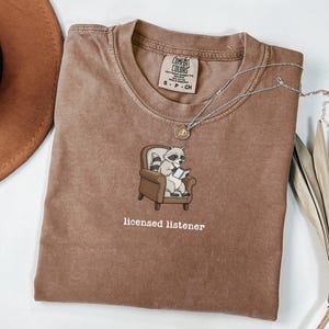 May include: A brown t-shirt with a graphic of a raccoon in a chair reading a book, with the text "licensed listener" below. A silver necklace with a small pendant is on the shirt.