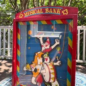 Pode incluir: Um cofre musical vintage de madeira vermelha com um painel frontal transparente. O cofre apresenta um design colorido de palhaços e as palavras "MUSICAL BANK" em letras douradas. O fundo é azul com confetes.