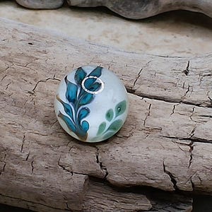 May include: A round glass bead featuring a translucent off-white base with a decorative pattern of dark teal, blue, and light green leaf-like shapes. The bead has a central hole surrounded by a silver-colored metal grommet for threading.