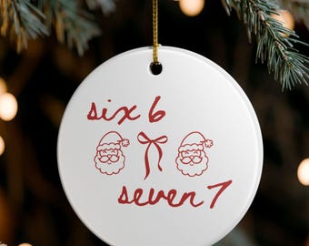 May include: A white, circular Christmas tree decoration with red text and illustrations. The ornament displays the numbers "six 6" and "seven 7", Santa Claus faces and a bow. A gold string is attached for hanging.