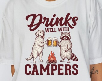 May include: White t-shirt with a graphic featuring a dog and raccoon toasting beer mugs over a campfire. The text reads "Drinks Well With Campers" in a burgundy script. A casual, comfortable tee for camping enthusiasts.