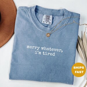 Op de afbeelding: Een blauw Comfort Colors T-shirt met de witte tekst "merry whatever, i'm tired". Een gouden ketting met een hanger ligt op het shirt. Een bruine hoed is zichtbaar in de linkerbovenhoek. Een gele cirkel met "SHIPS FAST" staat rechtsonder.