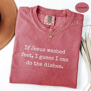 May include: A crimson Comfort Colors t-shirt with the text "If Jesus washed feet, I guess I can do the dishes." A gold pendant necklace is draped across the shirt. The shirt has a crew neck and short sleeves.