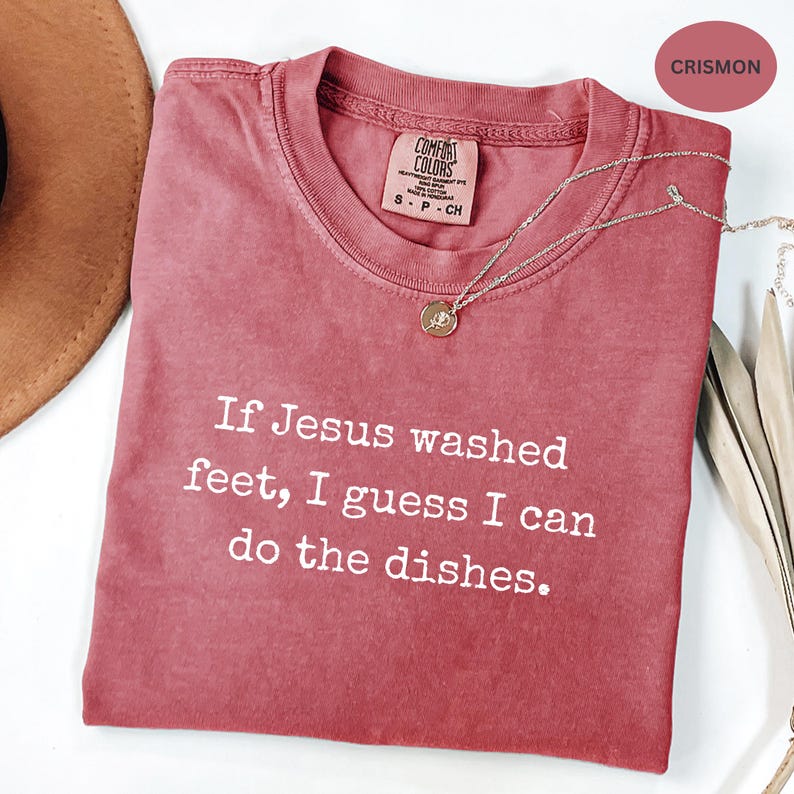 May include: A crimson Comfort Colors t-shirt with the text "If Jesus washed feet, I guess I can do the dishes." A gold pendant necklace is draped across the shirt. The shirt has a crew neck and short sleeves.