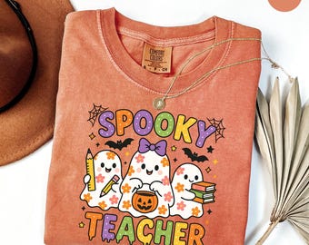 Puede incluir: Una camiseta Comfort Colors color ñame con las palabras "Spooky Teacher" y un gráfico de tres personajes fantasma. Los fantasmas sostienen un lápiz, una calabaza y libros. También se ve un collar dorado y un sombrero marrón.