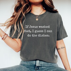 May include: Dark grey t-shirt featuring the quote: "If Jesus washed feet, I guess I can do the dishes." The shirt has a relaxed fit and a crew neck. The text is in a simple, sans-serif font.