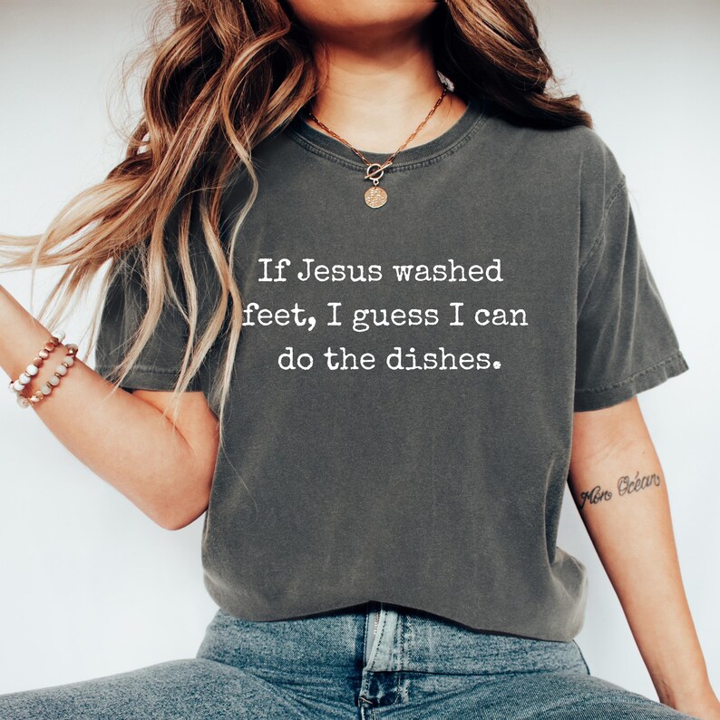 May include: Dark grey t-shirt featuring the quote: "If Jesus washed feet, I guess I can do the dishes." The shirt has a relaxed fit and a crew neck. The text is in a simple, sans-serif font.