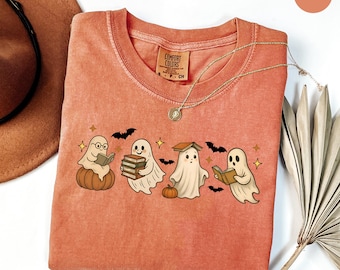 Op de afbeelding: Een koraalkleurig Comfort Colors T-shirt met een afbeelding van vier stripgeesten die boeken lezen, met vleermuizen en sterren. De geesten zitten op pompoenen. Een gouden ketting is over het shirt gedrapeerd. Het shirt heeft de tekst "COMFORT COLORS".