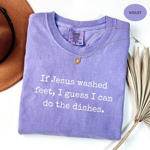 May include: A violet Comfort Colors t-shirt with the text "If Jesus washed feet, I guess I can do the dishes." A gold necklace with a pendant rests on the shirt. A brown hat and dried plant are in the background.