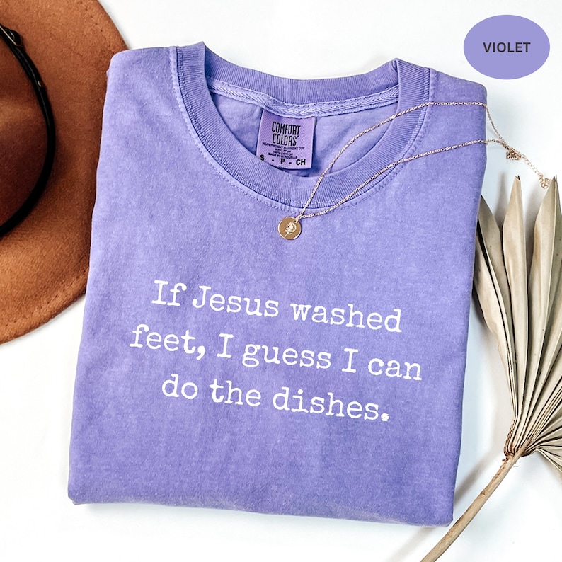 May include: A violet Comfort Colors t-shirt with the text "If Jesus washed feet, I guess I can do the dishes." A gold necklace with a pendant rests on the shirt. A brown hat and dried plant are in the background.