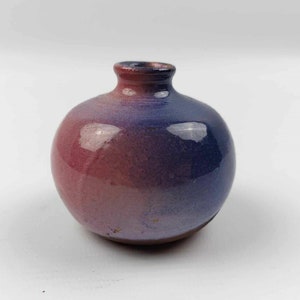 Pode incluir: Um pequeno vaso de cerâmica redondo com um pescoço curto. O vaso tem um esquema de cores gradiente, passando do vermelho profundo na base para o azul vibrante no topo. O acabamento brilhante reflete a luz, destacando a superfície lisa.