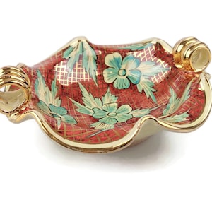 Peut inclure: Un plat décoratif en céramique avec un bord festonné et deux poignées dorées. Le plat présente un fond rouge avec un motif quadrillé et des motifs floraux et de feuilles turquoise. Le bord et les poignées sont dorés.