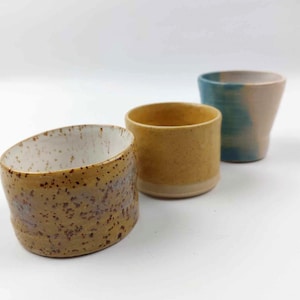 May include: Three handmade ceramic cups. The first is light brown with dark speckles and a white interior. The second is yellow with a light brown base. The third is blue and tan.