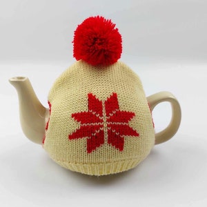 Può includere: Una teiera color crema con una coperta per tè lavorata a maglia. La coperta presenta un motivo a fiocco di neve rosso e un pompon rosso sulla parte superiore. La teiera ha un beccuccio e un manico curvi.