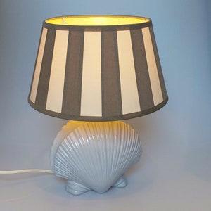 Può includere: Una lampada a forma di conchiglia bianca sostiene un paralume a strisce verticali grigie e bianche alternate. La lampada è accesa, emettendo un bagliore caldo. Il cavo è bianco.